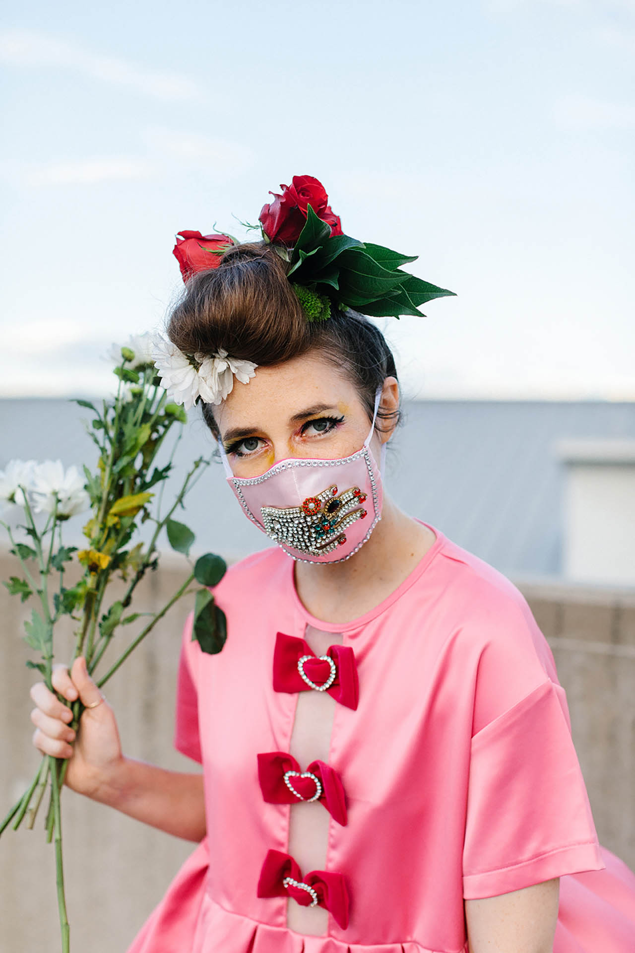 DFW editorial photos; a woman wearing a pink shirt and a pink mask covered in a rhinestones that creates a bejeweled hand on the mask. She is holding a small bouquet of white flowers while standing outside in an urban setting.