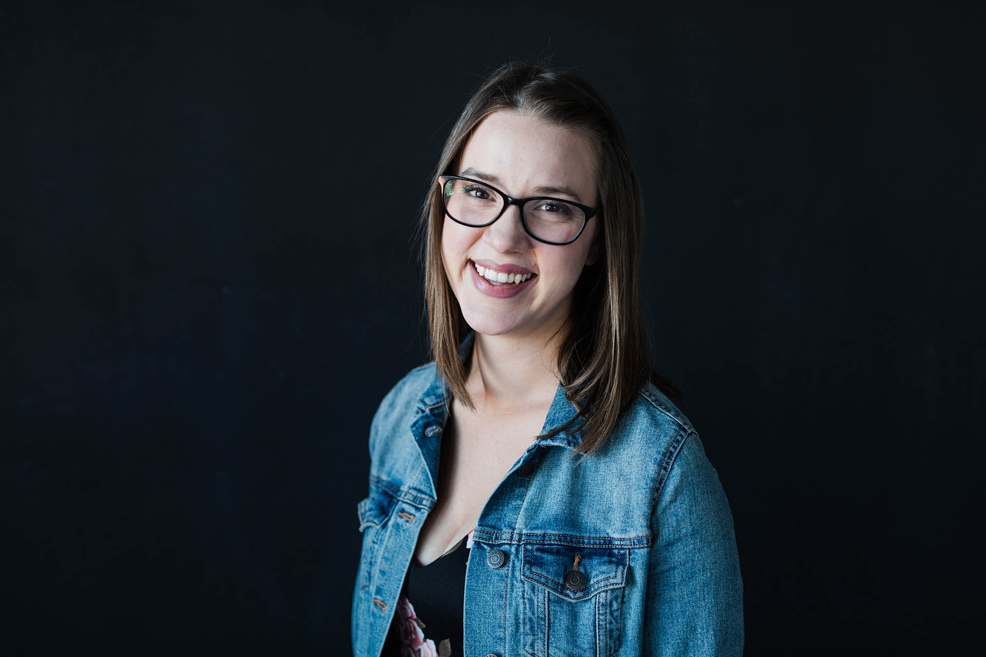 Fort Worth headshot photography; photo of a woman in a black top and denim jacket and glasses smiling in front of a black background