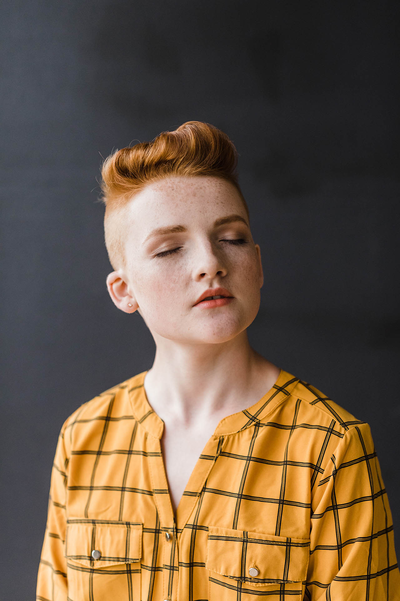 Fashion editorial photography; close up of a teenager wearing makeup and an orange and black striped shirt with closed eyes and slightly parted lips in front of a dark background