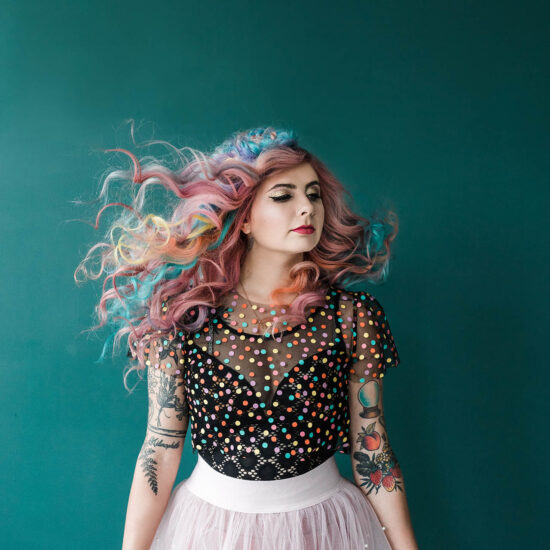 Dallas Commercial Photography; a woman with flowing multi-colored hair wearing elaborate makeup and a colorful, polka dotted top looking off into the distnace in front of a blue background