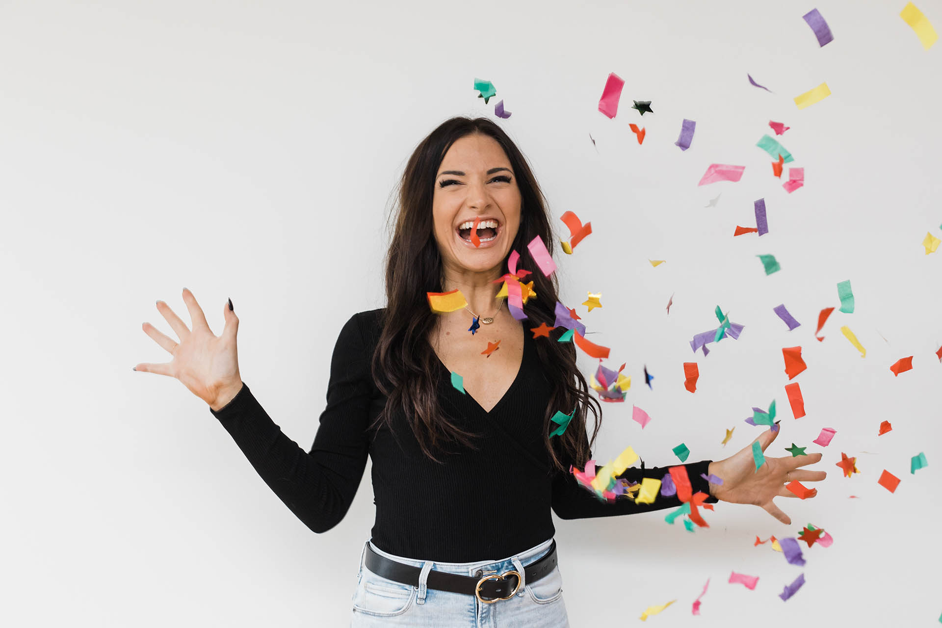 Caucasian woman in a black shirt and jeans smiling joyously after tossing confetti into the air in front of a white backdrop