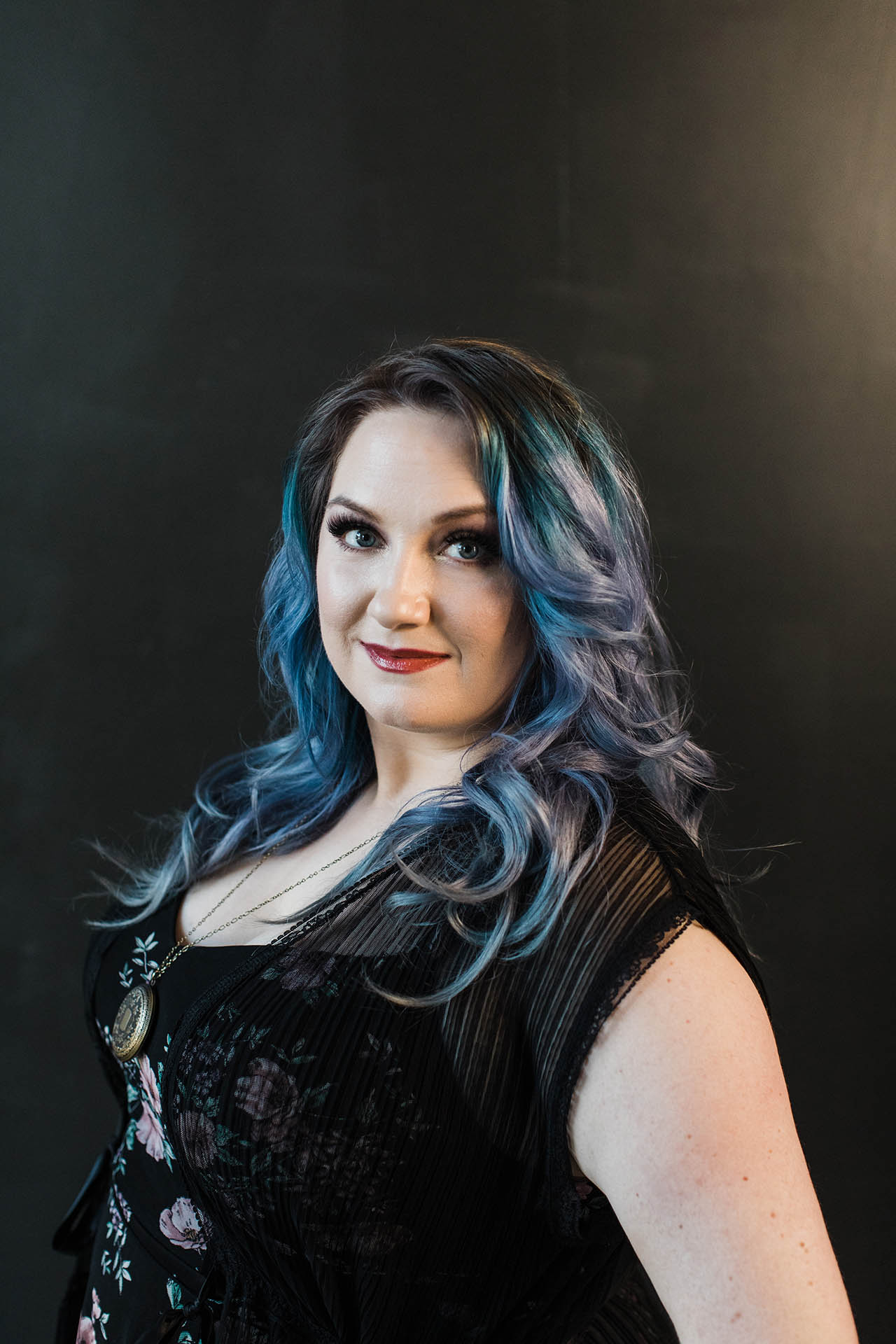 Head shot photographer Dallas; photo of a woman in a black top with colorful blue and purple hair smiling in front of a black backdrop.