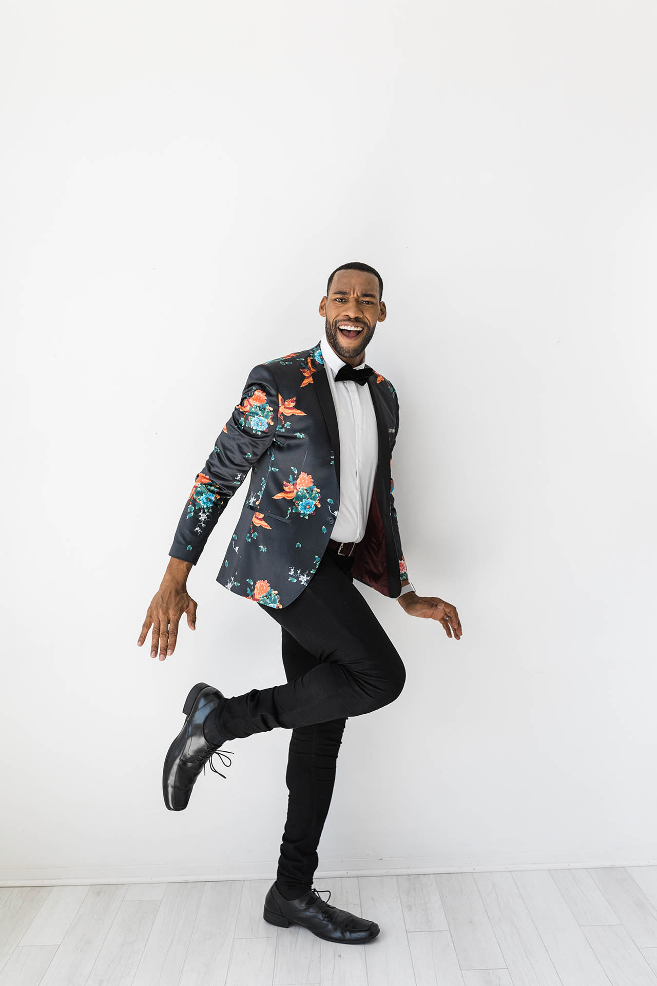 Men's fashion photos; detail shot of an African American male's stylish black and floral suit jacket. He is posed energetically with a smile in front of a white background.