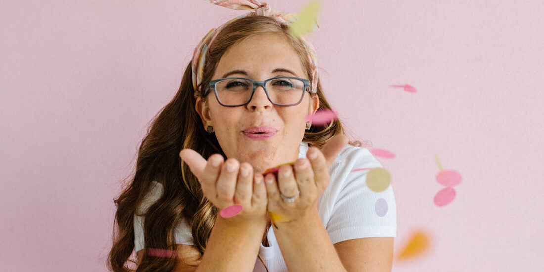Dallas Commercial Photography; photo of a woman wearing a colorful headband, white shirt, and glasses blowing colorful confetti circles into the air in front of a light pink background