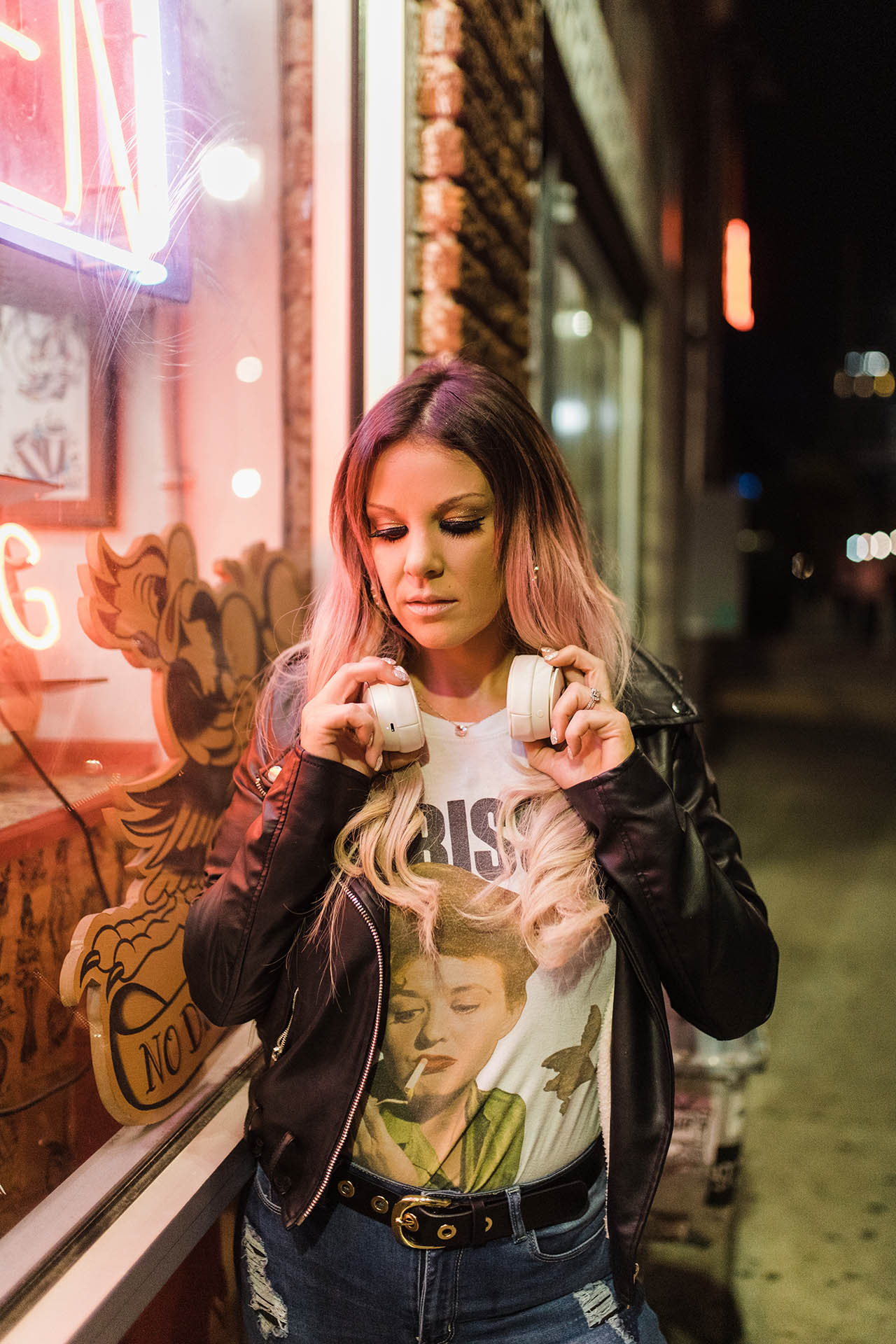 DFW branding photos; a woman wearing elaborate makeup, a black leather jacket, and a white graphic shirt looking down and holding the headphones around her neck in a downtown city setting