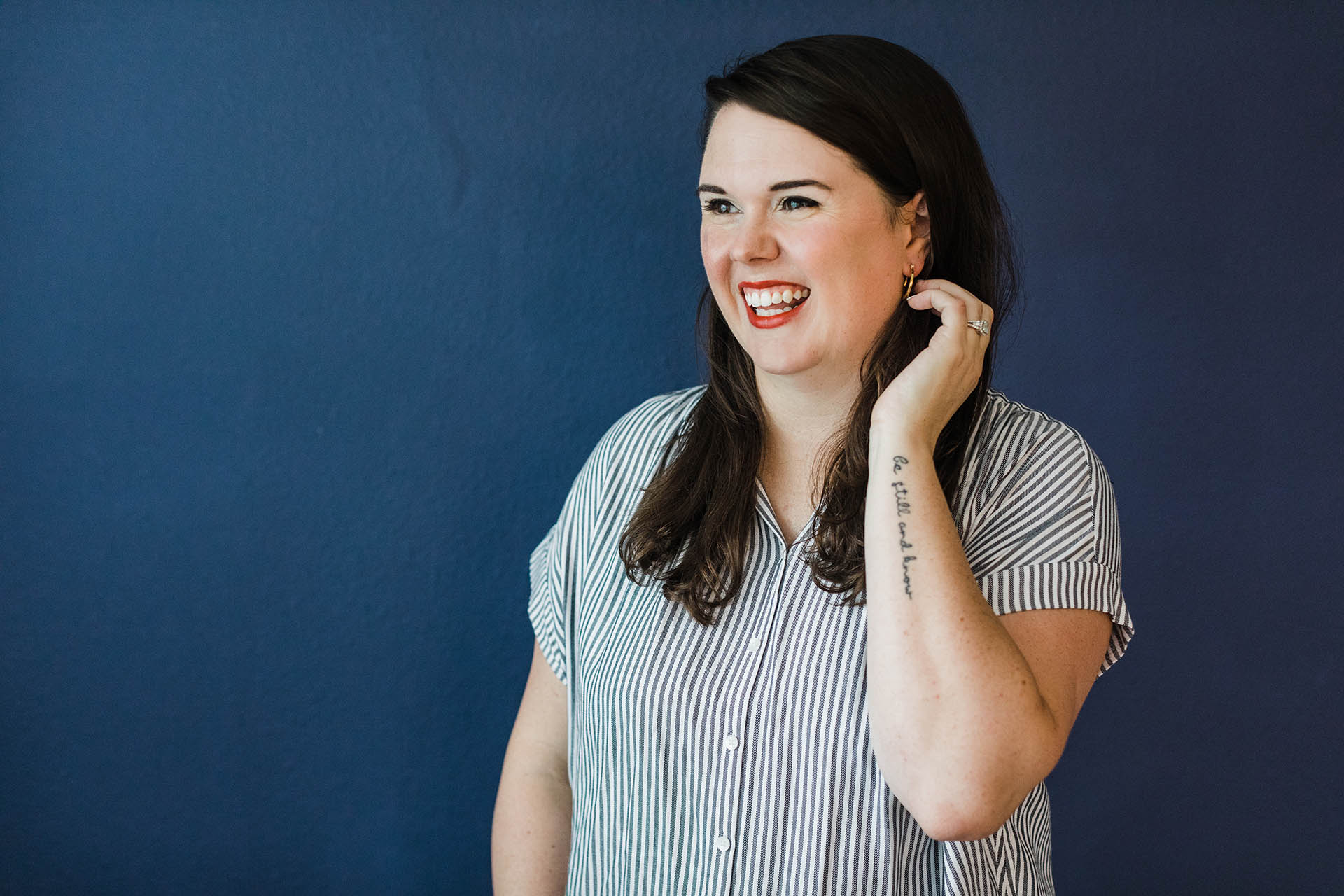 Corporate photographer DFW; a woman wearing a blue and white striped shirt smiling and looking off into the distance in front of a blue background. Her left arm is reaching up towards her face and her forearm tattoo reads "Be still and know."
