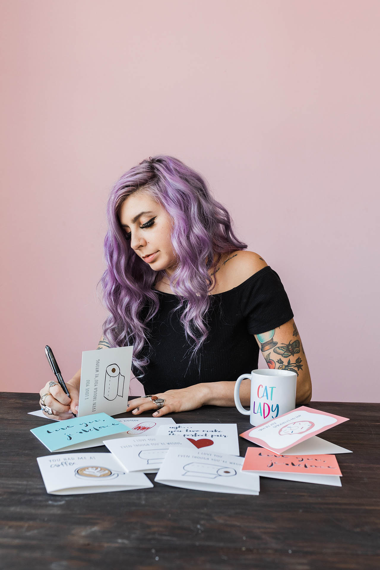 Dallas branding photographer; a woman with purple hair and tattoos wearing a black top and writing in a card in front of a light pink background
