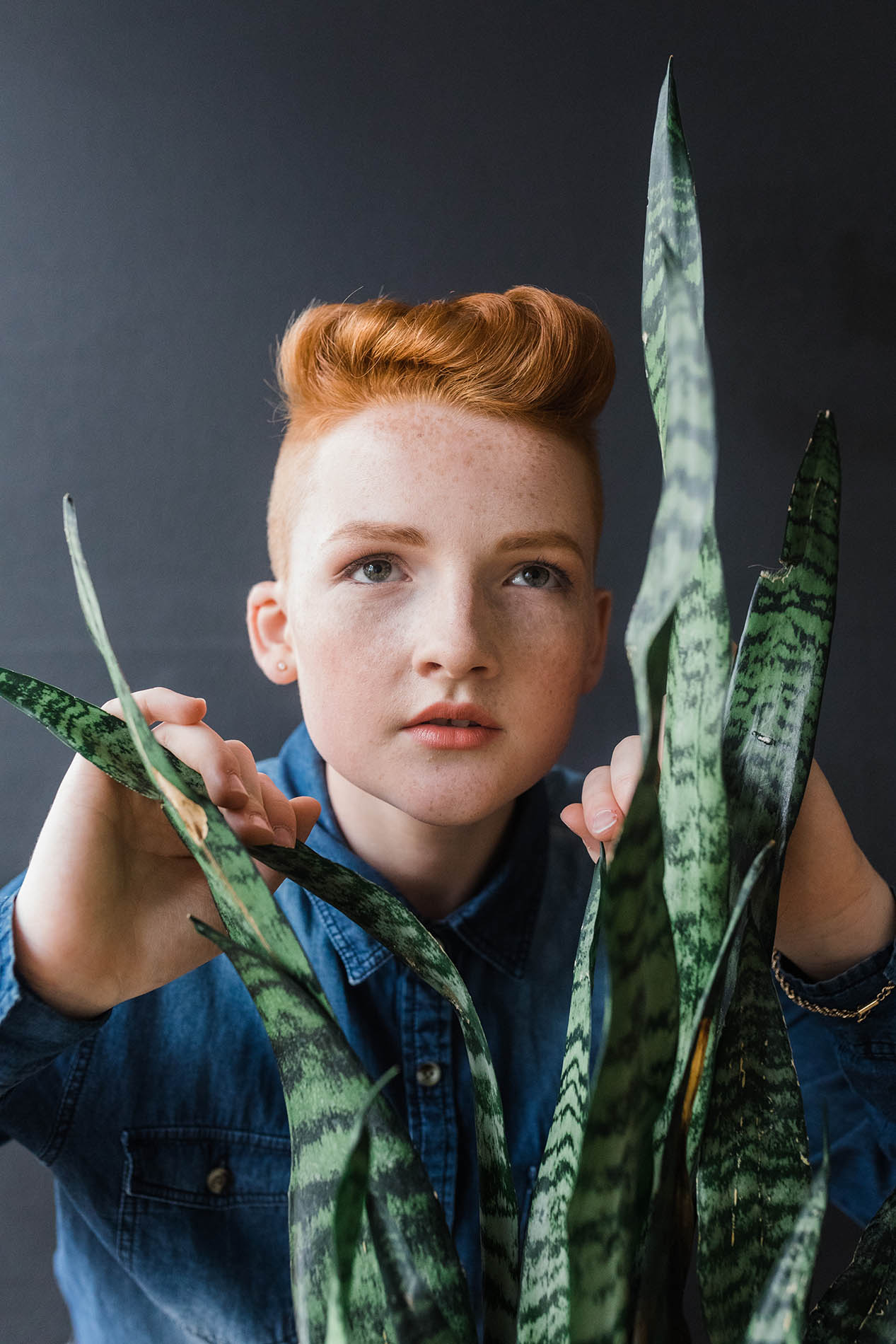 Dallas fashion photography; a young teen wearing a blue shirt, makeup, and an earring looking with determination through a plant in front of a dark background