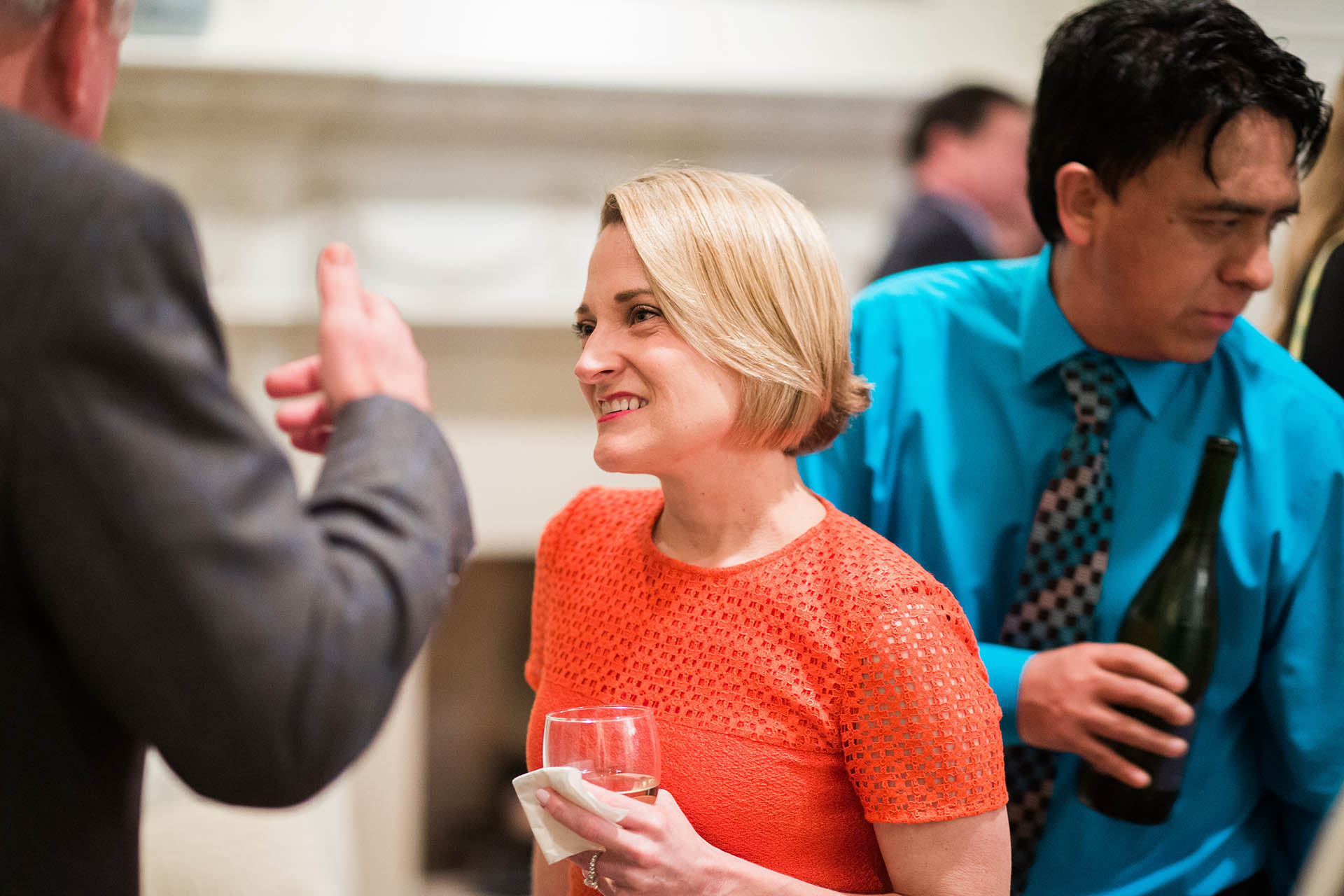 Event photography DFW candid photo of a woman in an orange dress smiling and conversing with another professional at a coporate event while holding a glass of wine.