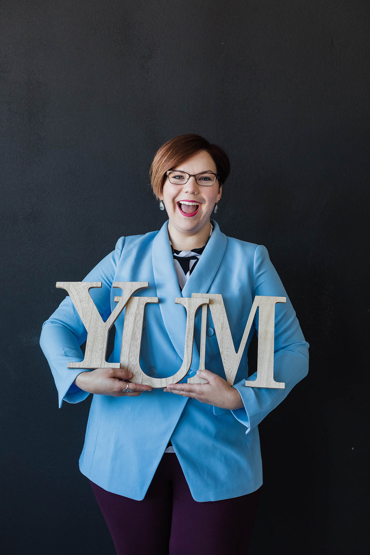Female corporate headshots; detail shot of a woman's lower face and torso where she's laughing, wearing a blue blazer, and holding a wooden cutout of the word "YUM" in front of a dark background