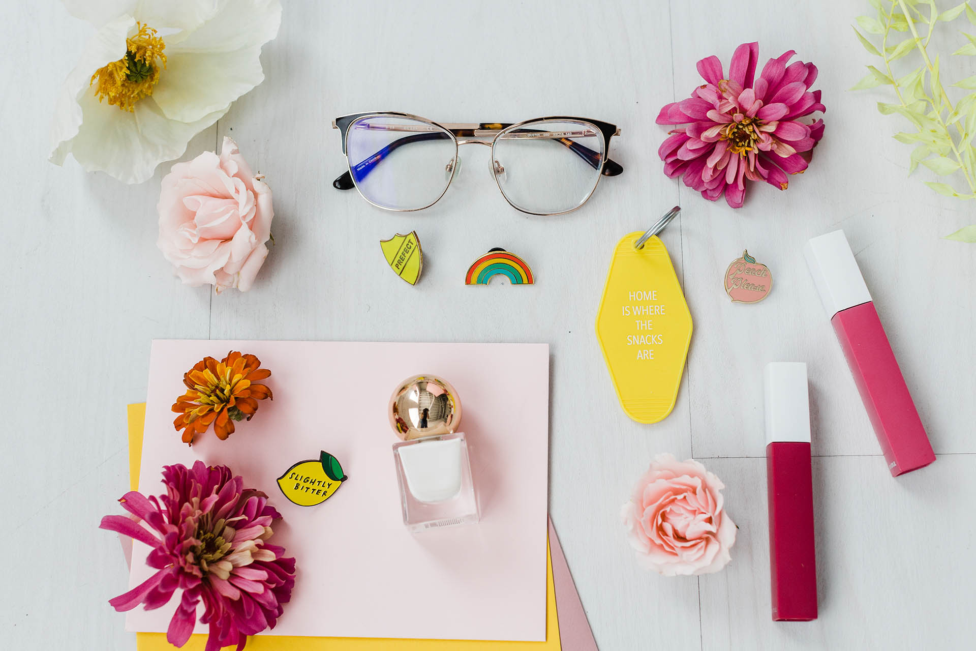 Flat lay photography; a detail photo of a flat lay of flowers, glasses, pins, cards, nail polish, and more all on a white, wooden floor