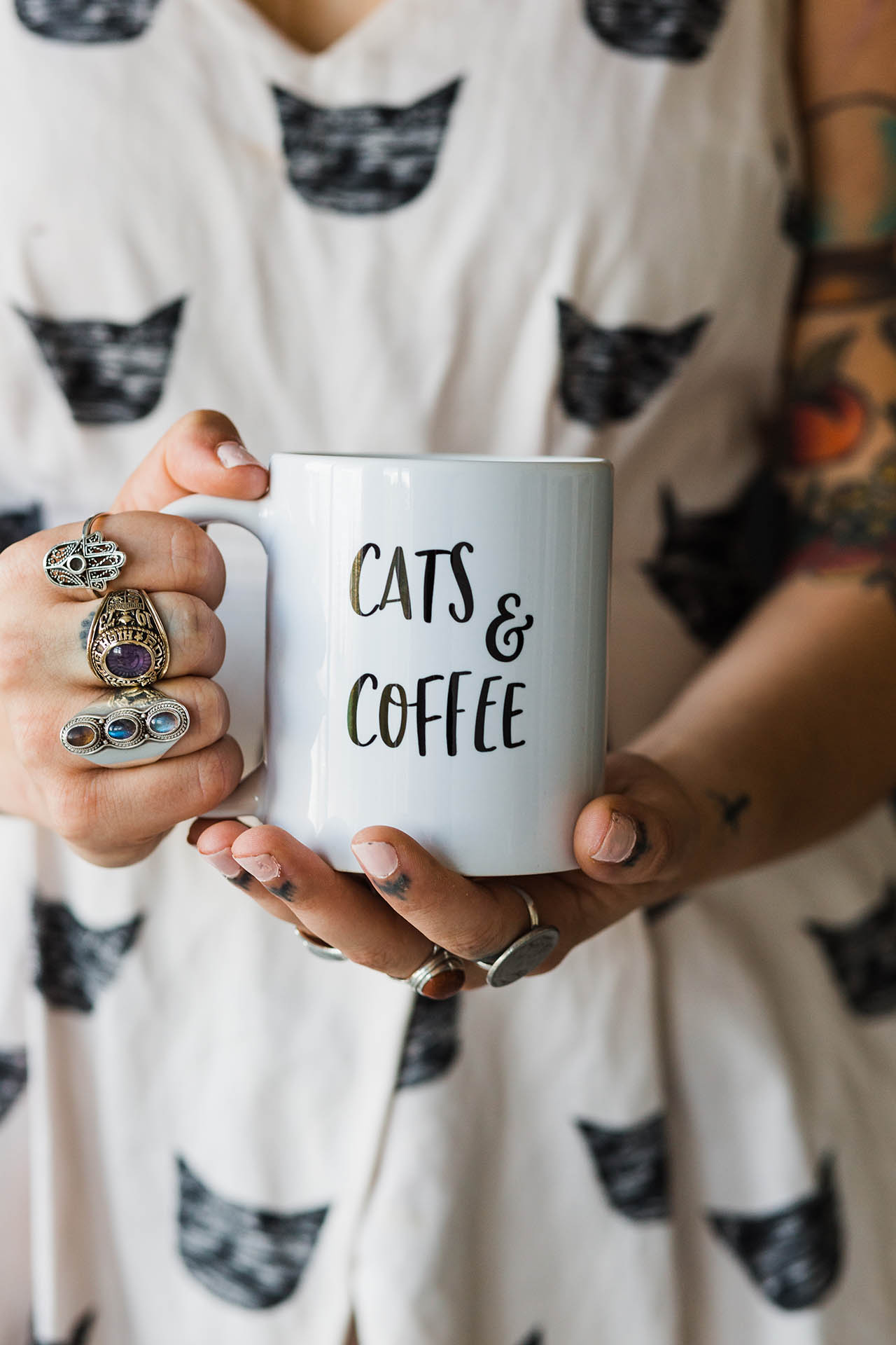Fun branding photographer; detail shot of two hands holding a coffee mug that reads "Cats & Coffee." The right hand has three different, detailed rings on the fingers.