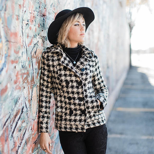 Lifestyle brand photographer Dallas; a woman wearing a black and white checkered tweed jacket, black pants, black shirt, silver necklace, and black hat looking thoughtfully into the distance while resting against a brick wall covered in graffiti