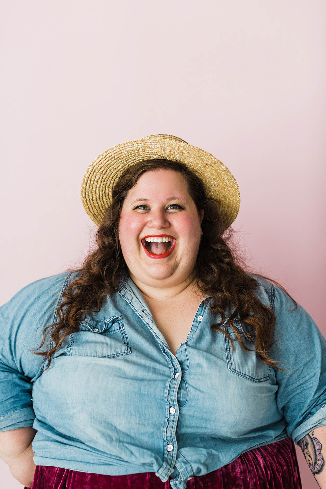 Plus size photography poses; a woman wearing a straw hat and a denim shirt smiling widely in front of a light pink background