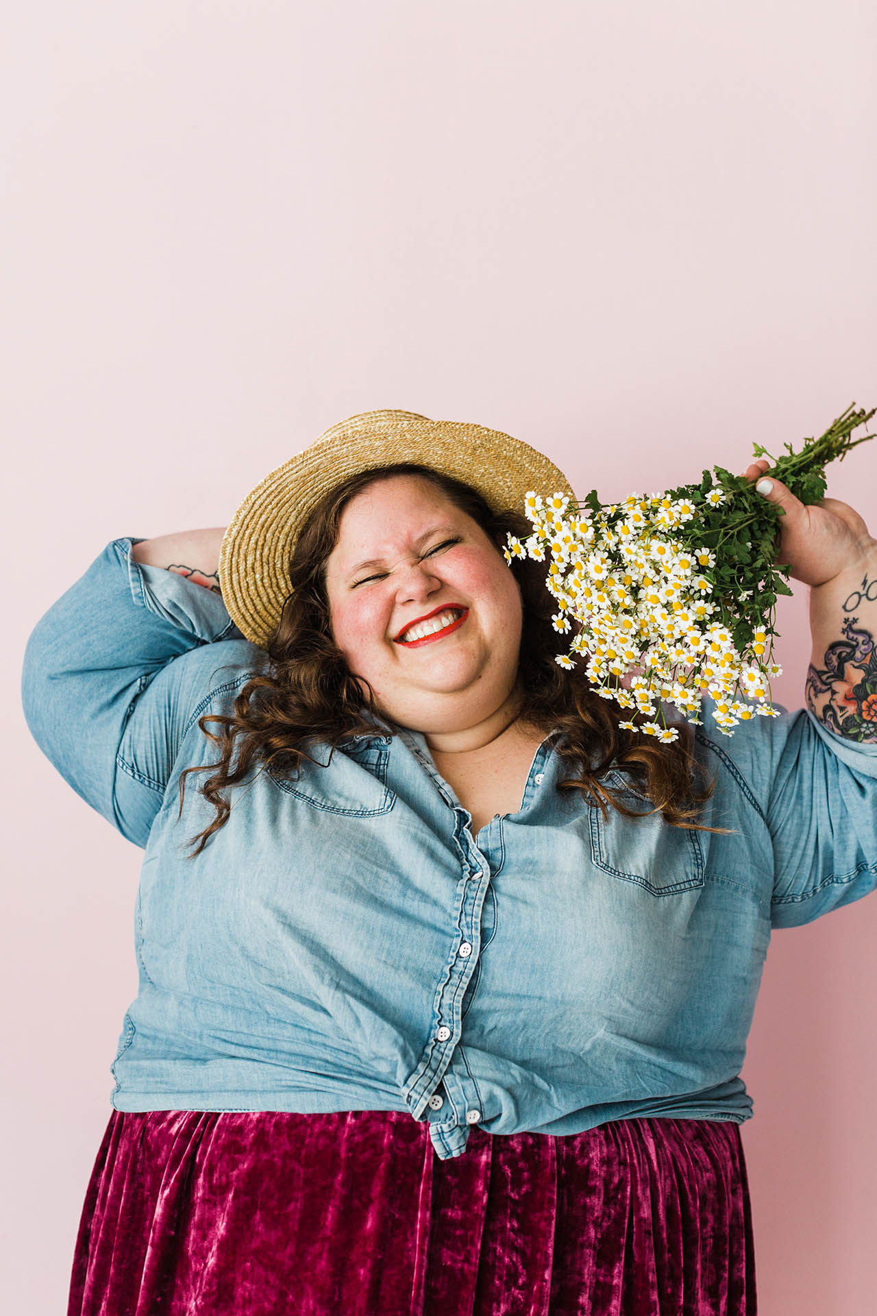 Plus size photography; a woman wearing a straw hat and a denim shirt smiling with closed eyes and holding a bouquet of flowers up to her face in front of a light pink background