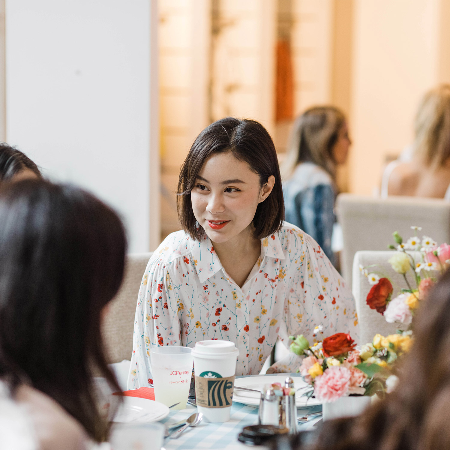 Candid moments event photos; candid shot of a woman are a business luncheon; she is wearing a floral dress shirt and is engaged with her fellow event guests while all sitting around a floral centerpiece on their table