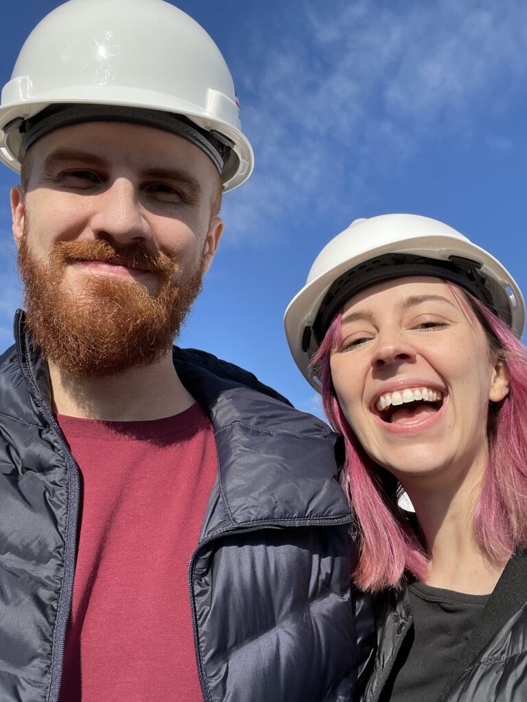 Photography couple wearing hard hats and jackets outside in front of the sky at a construction site