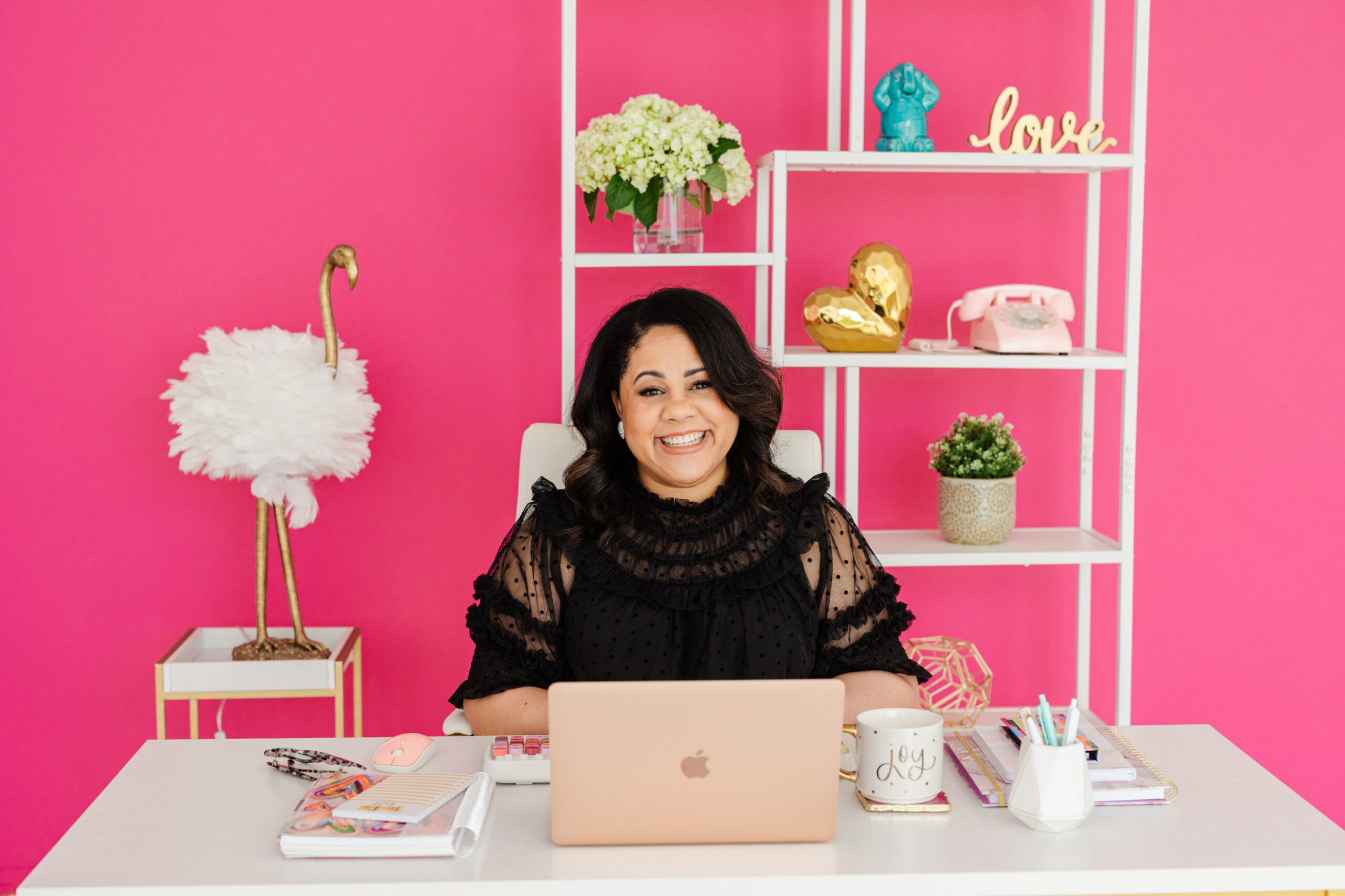 a woman having a professional business branding session she is sitting at a white desk in a white chair and behind her is a pink wall and a shelf with various objects