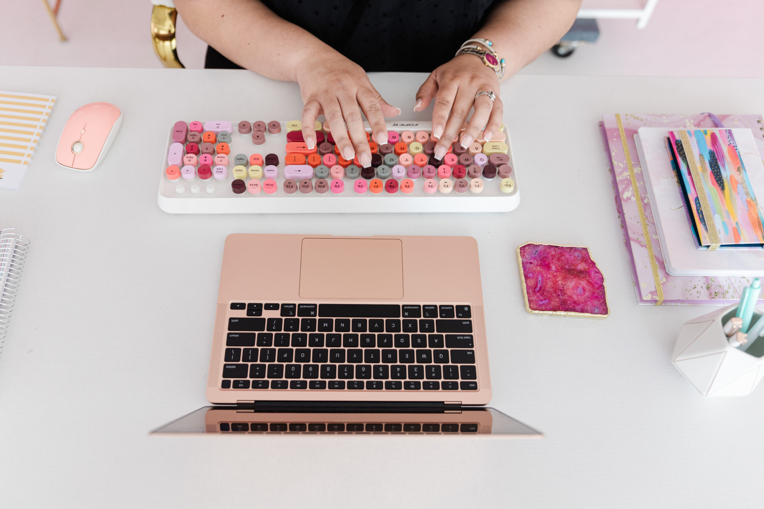 a woman typing on a colorful wireless keyboard also on the desk in a laptop, notebook, and pens
