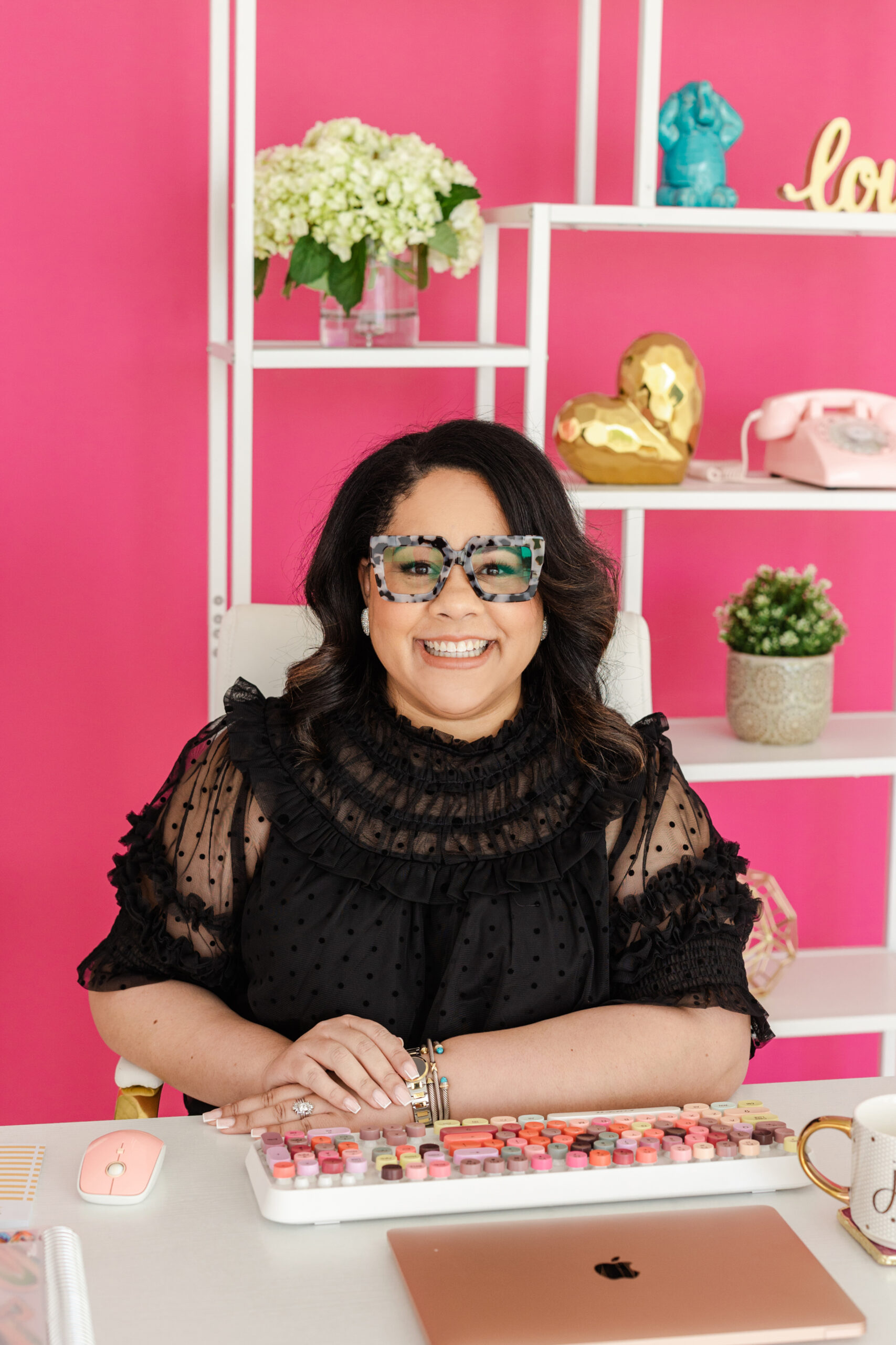 Jennifer from Joy of Work having a professional branding session in a studio behind her is a pink wall filled with different objects and in front of her is colorful keyboard and desk