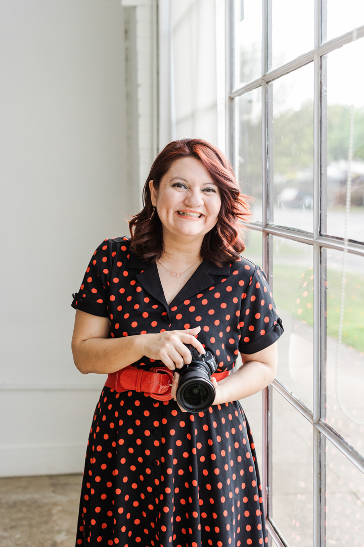 Colorful Dallas Headshot Photographer; photo of a woman in a black dress with red polka dots smiling and holding a camera in front of a window and a white wall.