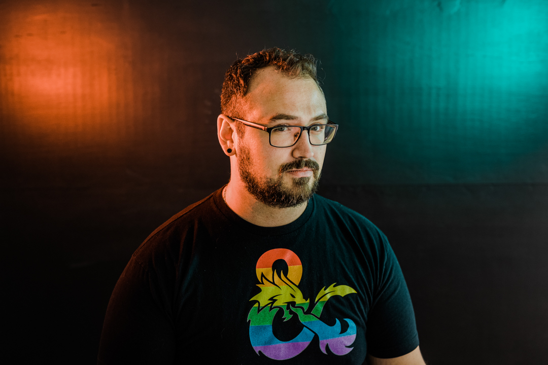 Colorful Dallas Headshot Photographer; photo of a man in a LGTBQ Dungeons and Dragons shirt and glasses looking coyly at the camera in front of a black background with orange and blue lights in the upper corners.