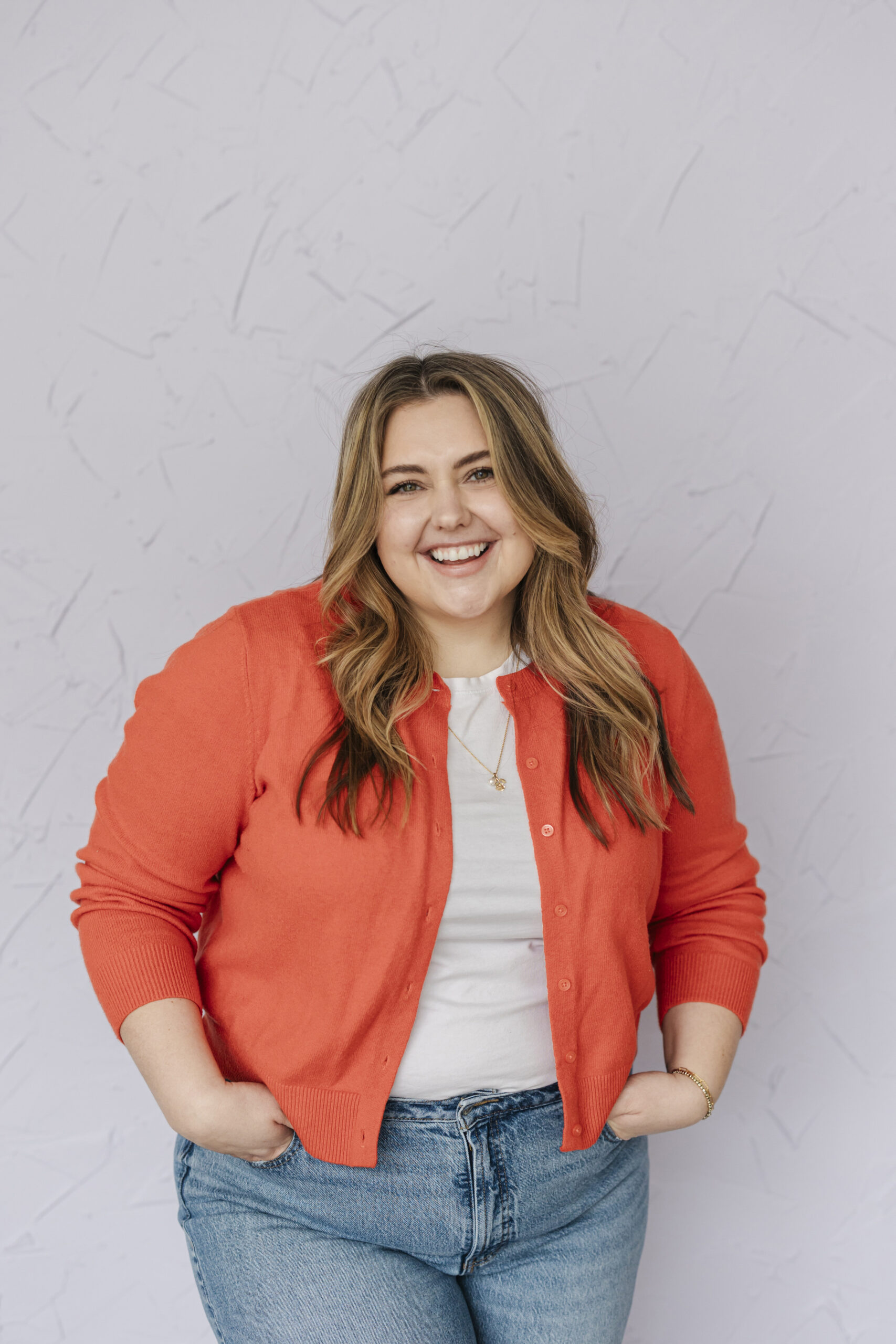 a woman wearing an orange cardigan standing next to a wall with her hands in her pockets taking lifestyle headshots 