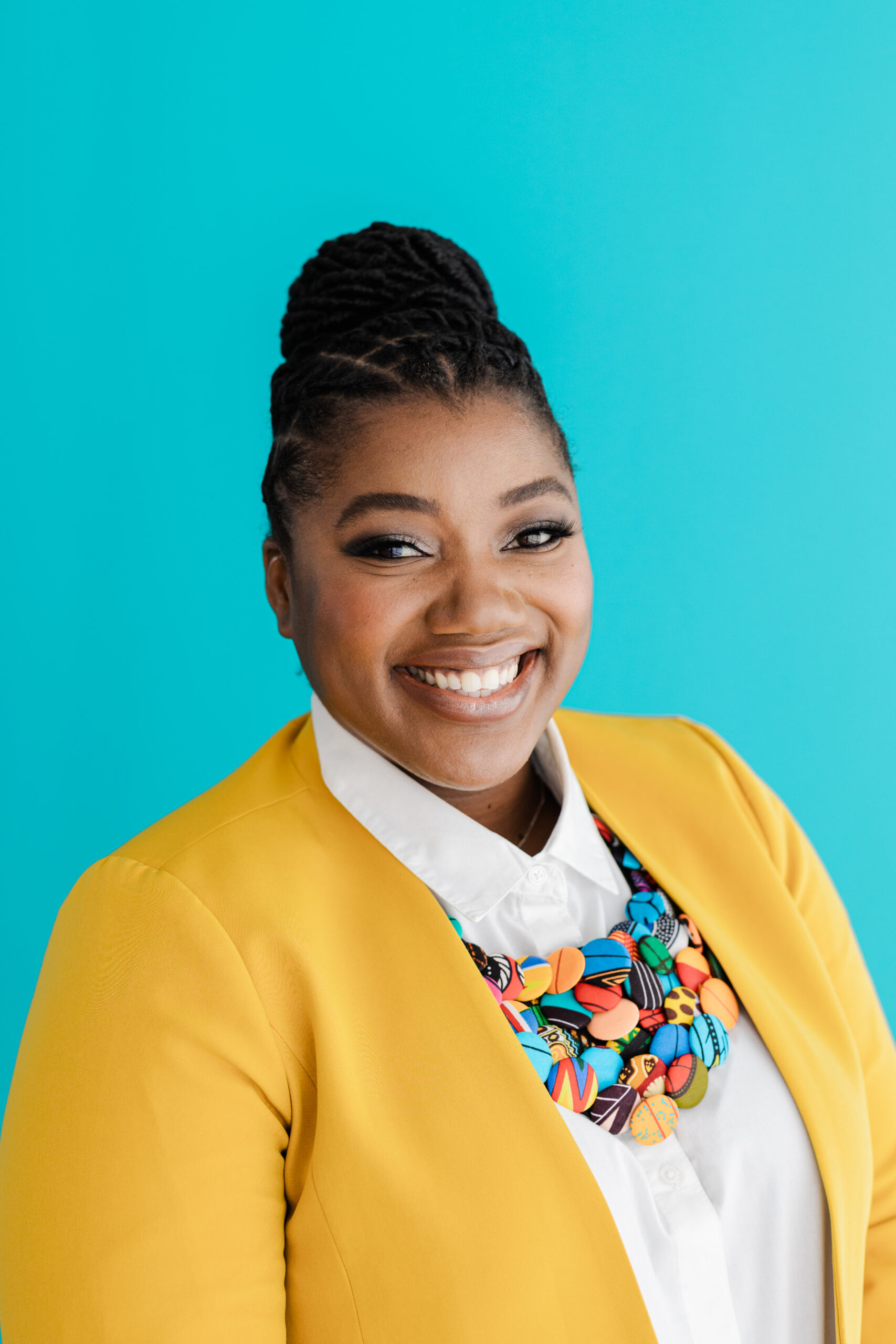 An African American woman smiles in front of a bright blue background. She is wearing a yellow jacket and a colorful necklace.
