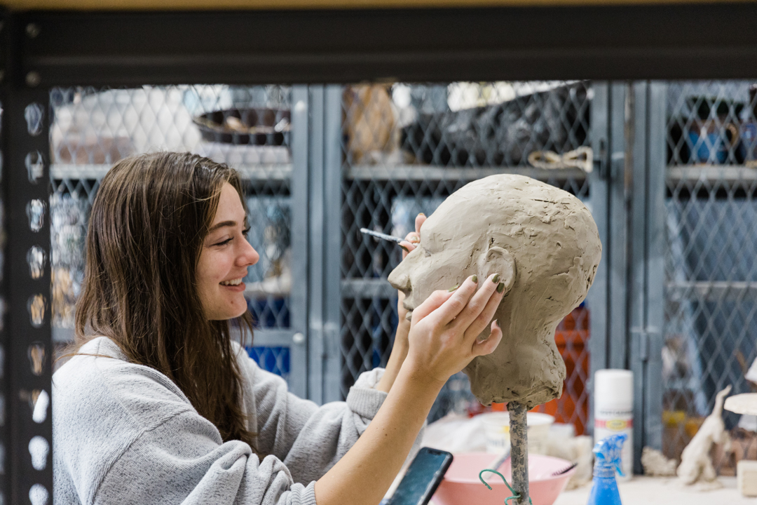 Dallas Artist Photographer; photo of a woman in a grey sweater smiling and working on a clay sculpture of a human head in an artist studio