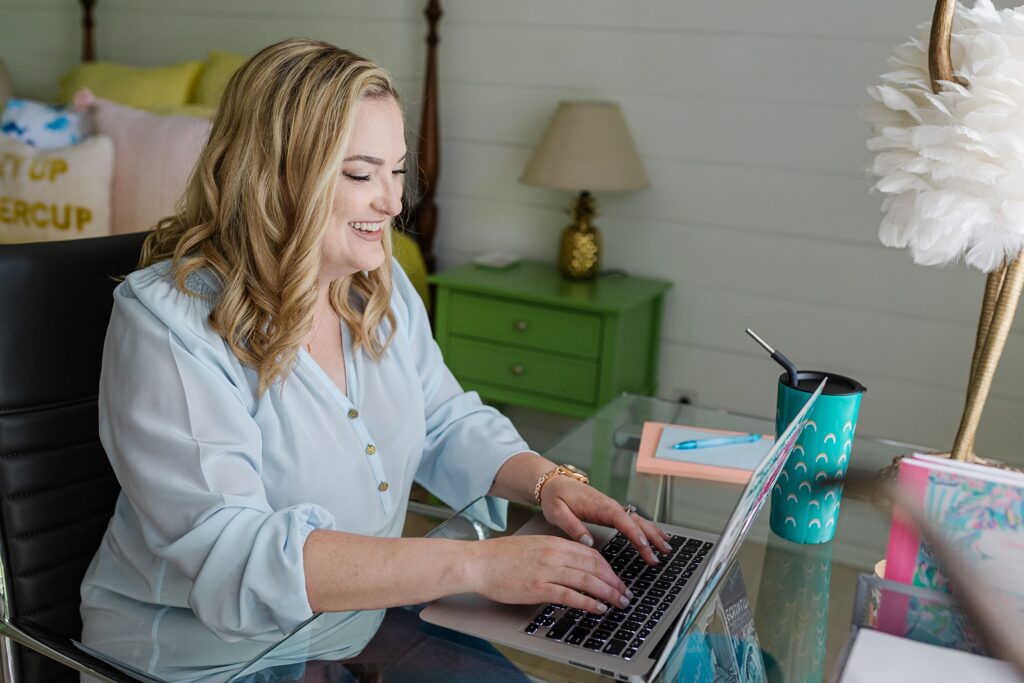 Blonde Caucasian woman smiling and typing on her laptop. She's wearin a light blue top and a gold watch and is sitting at a glass desk that has her laptop, a notebook, a thermos, and other decorations resting on top. The background is a bedroom setting with a green nightside table, a gold lamp, and a bed covered in decorative pillows.