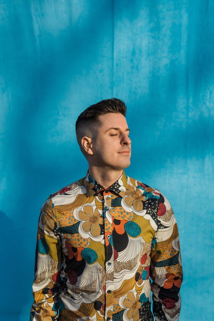 Caucasian male facing towards the sun with his eyes closed in front of a blue background. He has short dark hair and is wearing a highly detailed/colorful floral dress shirt.