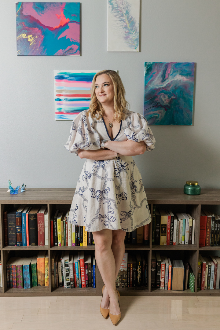 Dallas Brand Photographer; woman in a white and blue dress and heels looking off to the side with a slight smile and crossed arms in front of filled bookshelves and a white wall with two paintings