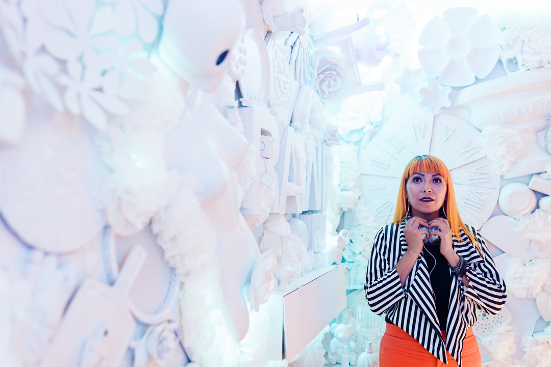 Dallas Brand Photographer; woman with dyed orange hair wearing bright orange pants, a black and white striped blouse, and silver headphones staring off into the distance in the midst of a all white art installation