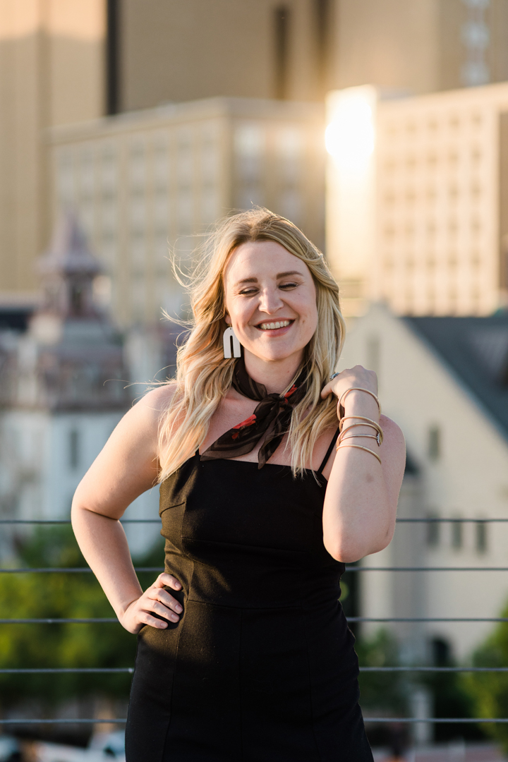 Dallas Brand Photographer; woman in a black dress and black handkerchief tied around her neck casually posing and smiling with closed eyes in an outdoor, downtown setting