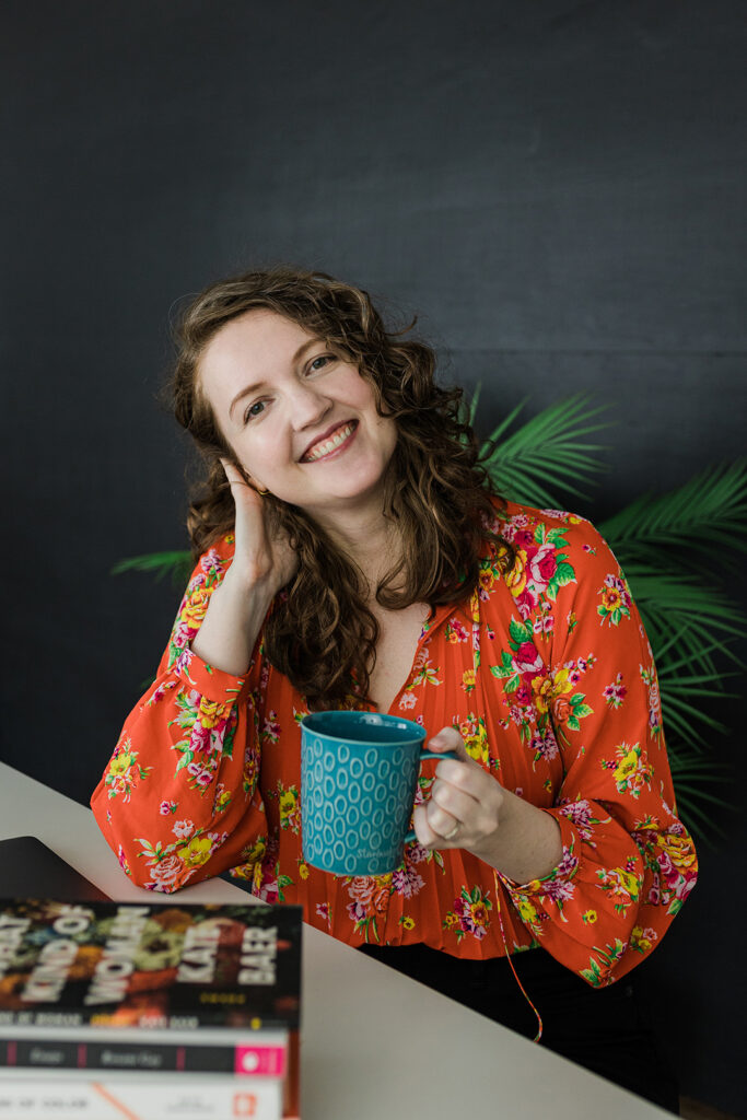 Dallas Brand Photographer; Caucasian woman wearing a floral blouse and holding a coffee cup while sitting a table with books on it
