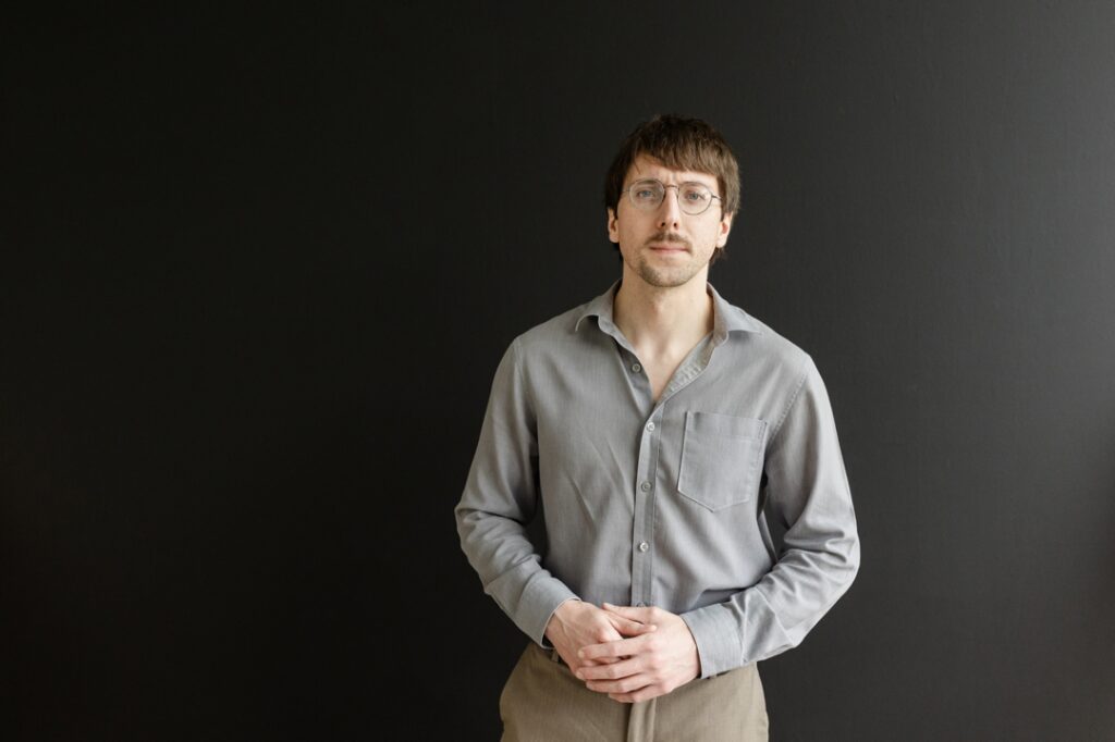 Caucasian man wearing glasses, a grey button down shirt, and brown pants, standing with folded hands in front of a dark backdrop