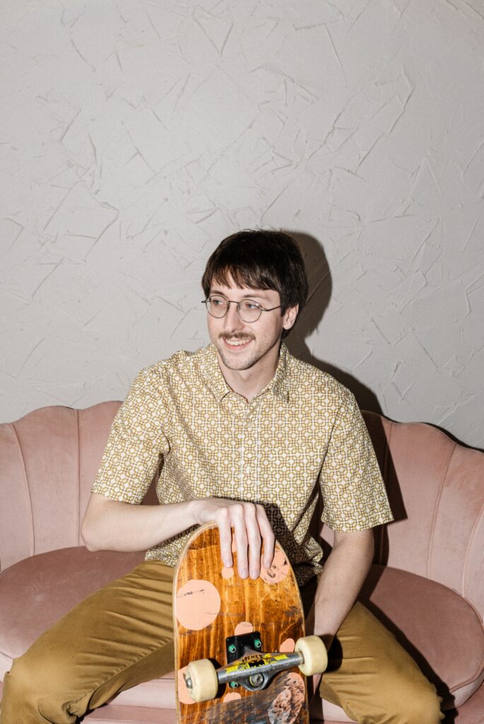 Caucasian man wearing glasses, a white and brown elaboratly patterned shirt, and brown pants, sitting on a salmon colored couch and holding a worn down skateboard while looking off to the side