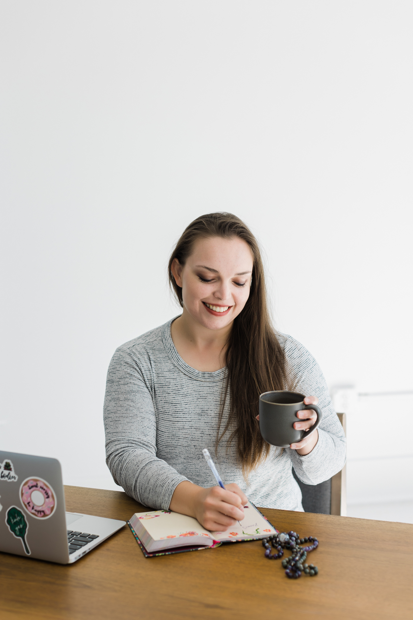 Dallas Brand Photographer; photo of a woman wearing a grey top smiling, holding a mug, and writing in a notebook on a desk with a laptop in front of a white background