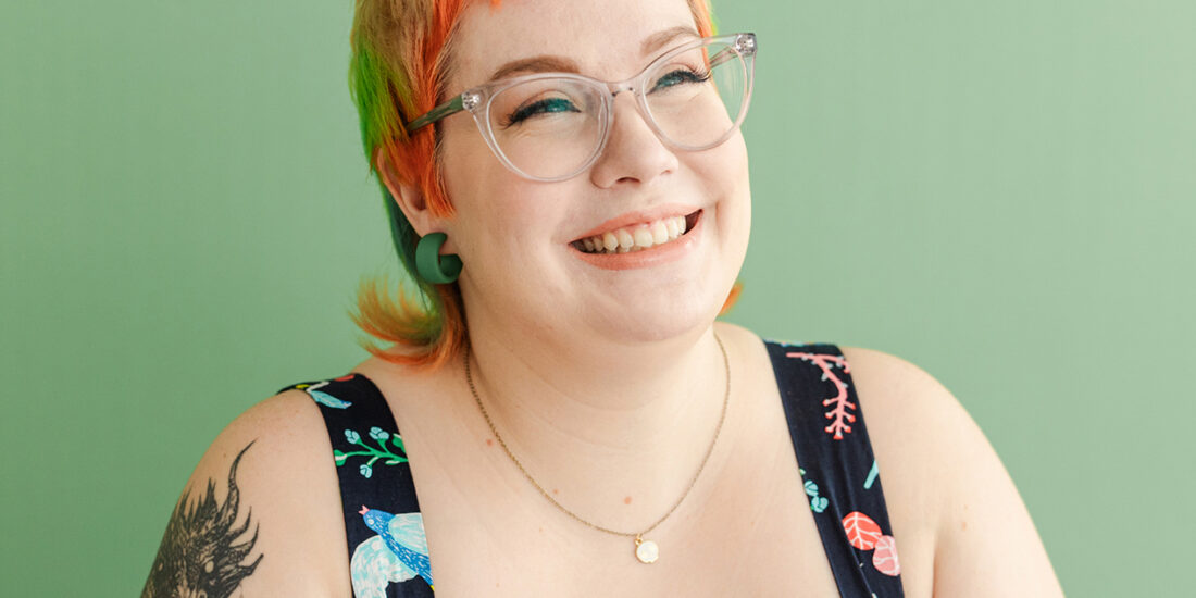 Dallas Branding Photographer: a woman smiling and looking off to the side in front of a green background. She's wearing clear glasses and a black dress decorated with colorful, detailed nature elements, and she also has colorful hair and tattoos.