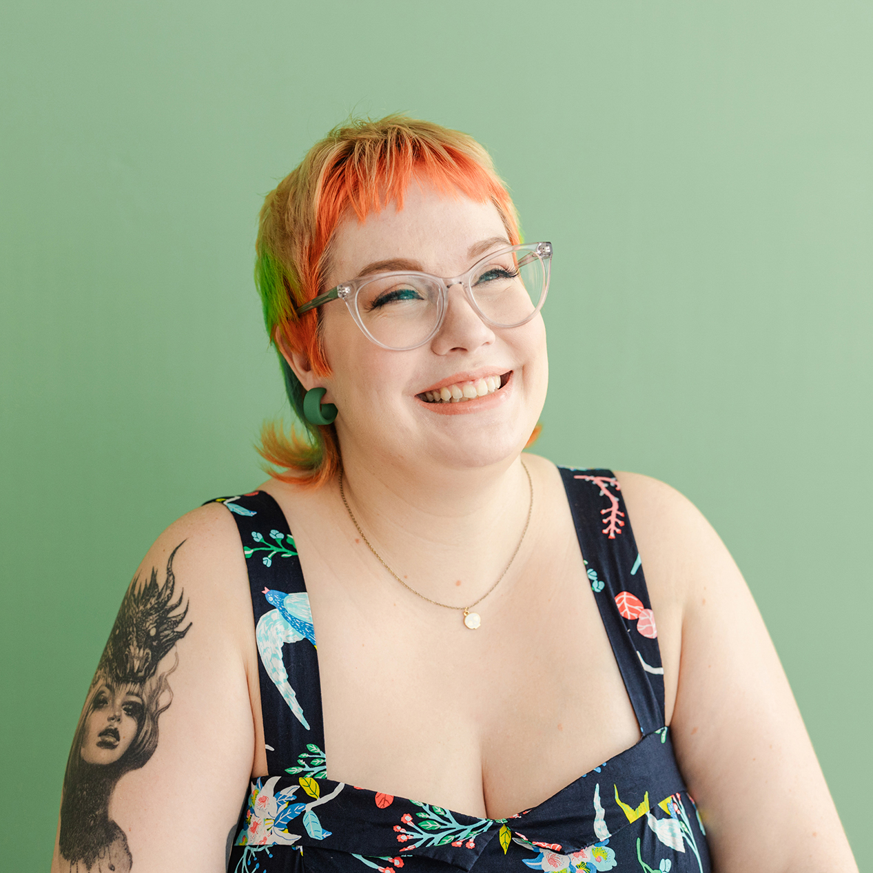 Dallas Branding Photographer: a woman smiling and looking off to the side in front of a green background. She's wearing clear glasses and a black dress decorated with colorful, detailed nature elements, and she also has colorful hair and tattoos.