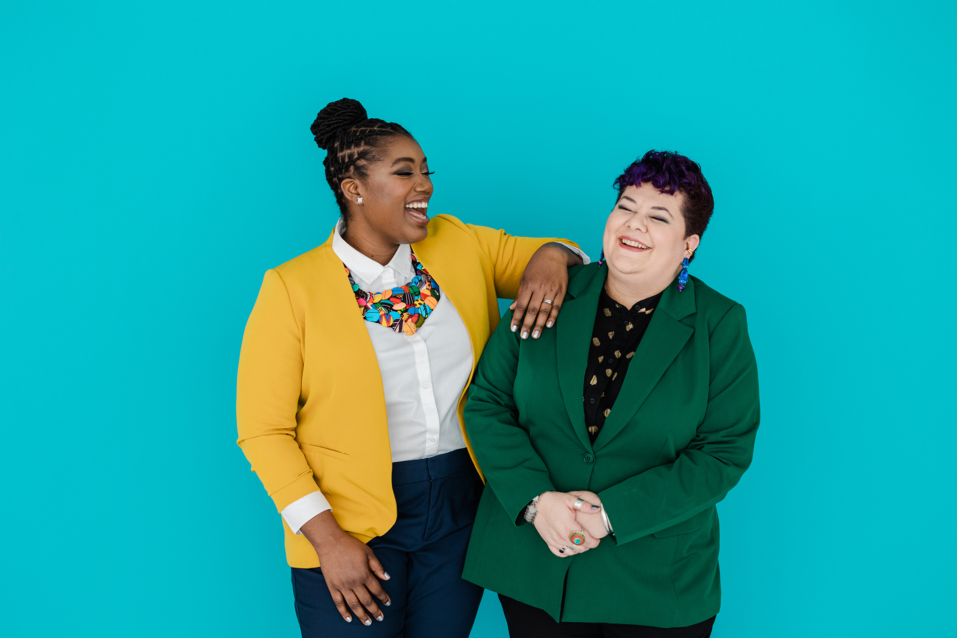 Dallas Branding Photographer: two women laughing and playfully posing together in front of a light blue background. Both are wearing colorful blazers and dress shirts and jewelry.