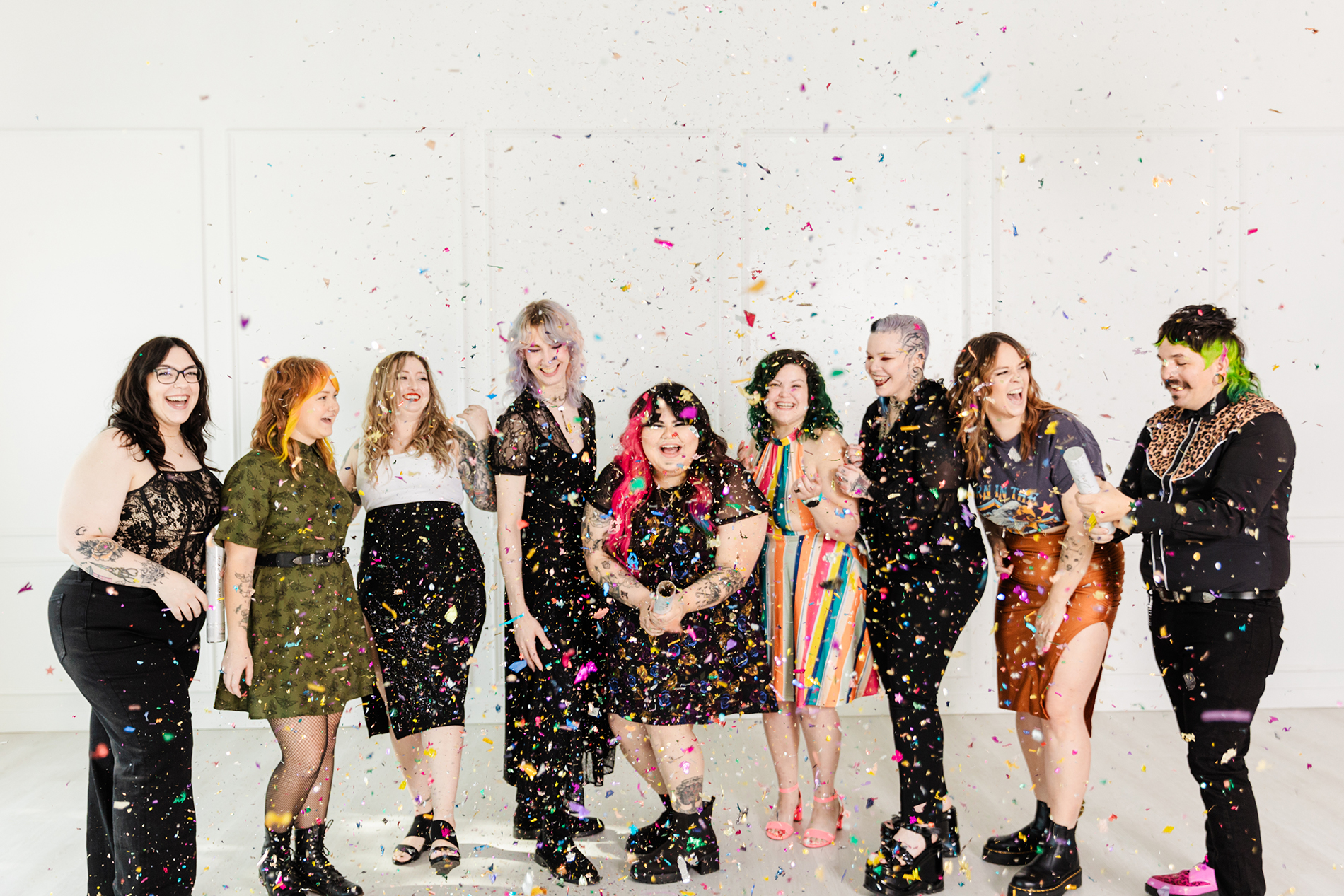 Dallas Branding Photographer: a business owner and her eight employees laughing and standing together in a line as they launch confetti poppers into the air. They're all wearing colorful, fun clothes and all have uniquely colored and styled hair.