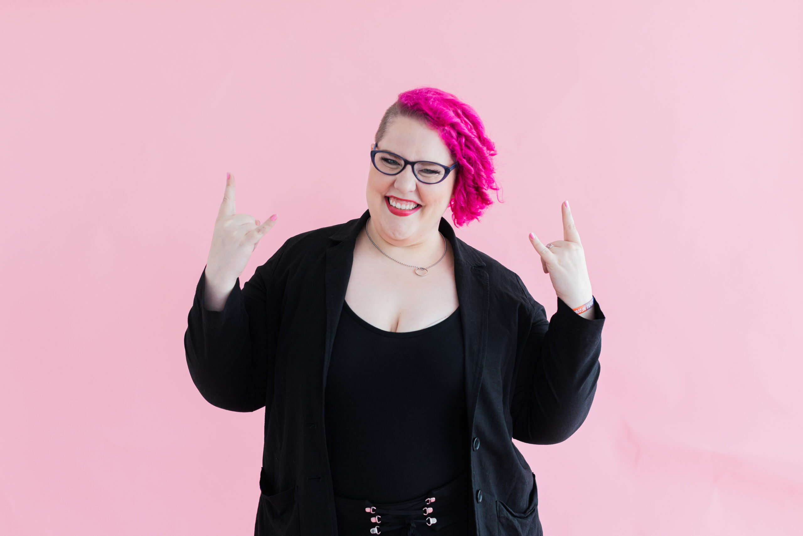 Dallas branding photographer; woman wearing all black and glasses with bright pink hair smiling and holding up rock n roll hands in front of a light pink background