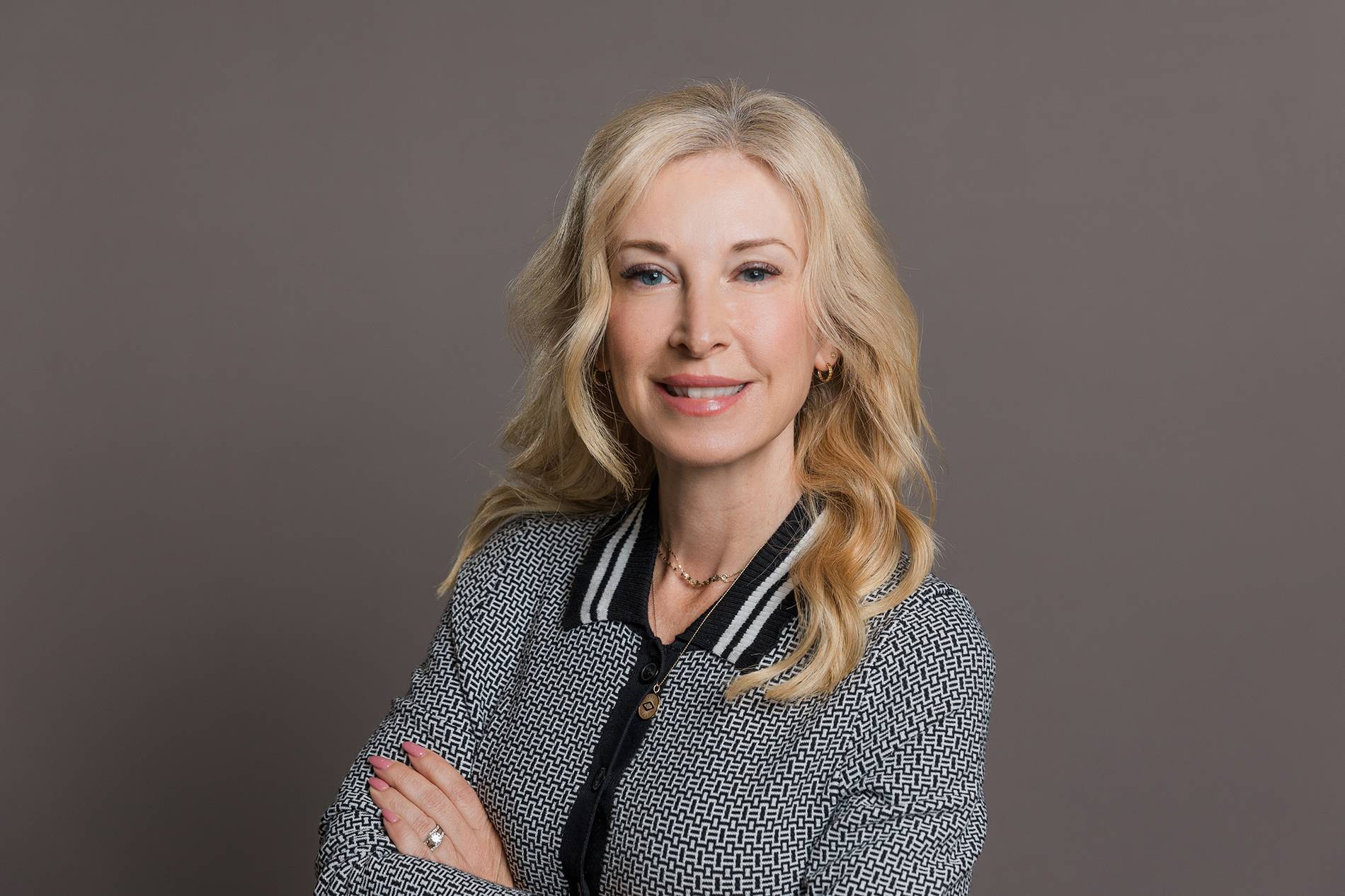 Dallas Corporate Head Shot Photographer: a woman smiling with folded arms in front of a dark background. She's wearing a black and white patterned top with a necklace.