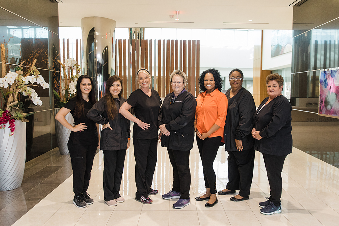 Dallas Corporate Lifestyle Photographer; seven women all in black (minus one woman wearing an orange shirt) all smiling and posing together in a fancy lobby