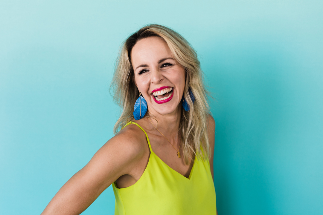 Dallas Editorial Brand Photographer; woman in a yellow green top and blue feather earrings laughing and looking to the side in front of a light blue background