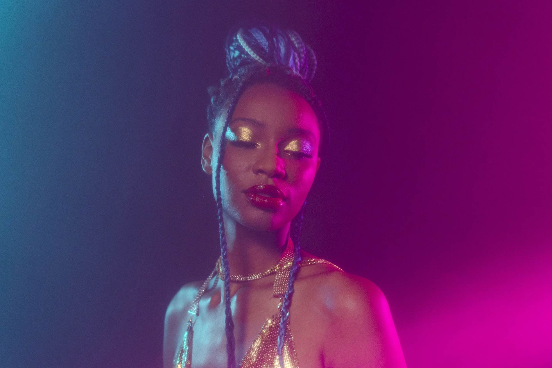 Dallas Editorial Photographer: a woman posing with closed eyes while bathed in blue and pink light. She has intricate gold makeup and jewelry alongside her intricate hairstyle.