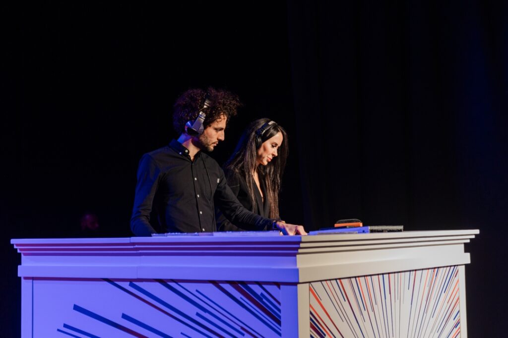 A man and woman DJ standing and working behind a colorful DJ booth while both wearing black and headphones
