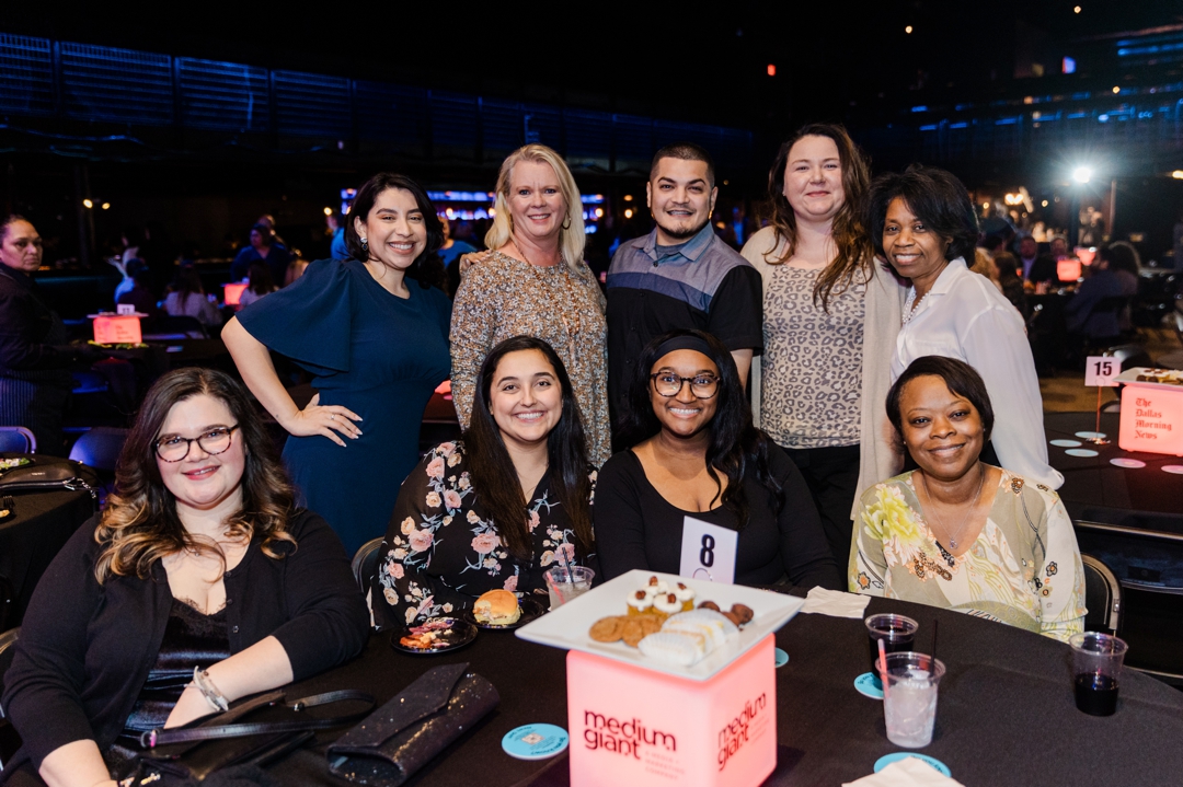 Dallas Event Photographer; Nine individuals posing and smiling in front of an event table