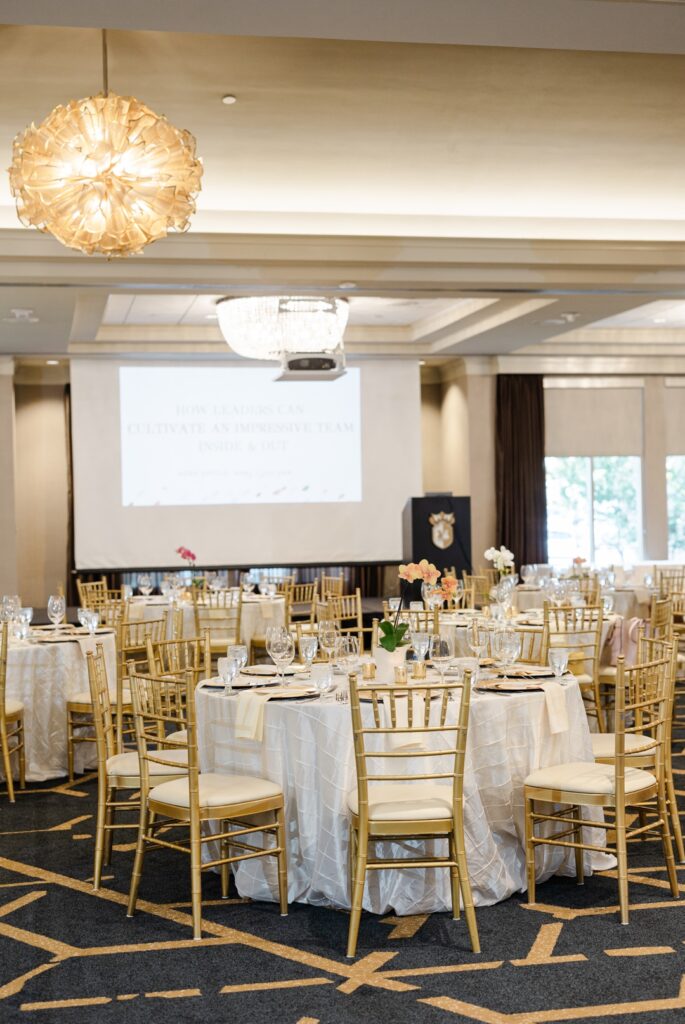 A detail photo of a table decorated with a tablecloth, glasses, plates, napkins, and a floral centerpiece. The table is surrounded by gold dining chairs wih cream cushions and the background contains more similar tables with a stage, podium, and projection screen.